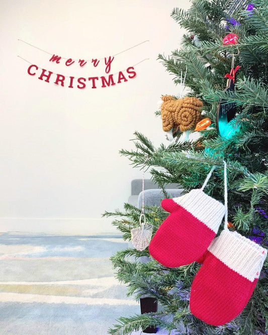 close up of christmas tree on the right with red mitten ornaments and rainbow lights. a hanging sign in background that says merry christmas in red
