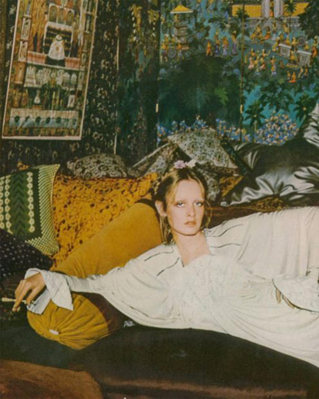 Twiggy the model laying on a sofa with a cigarette in her hand, surrounded by yellow and green floral pillows. A floral printed blue and green wall paper on the walls