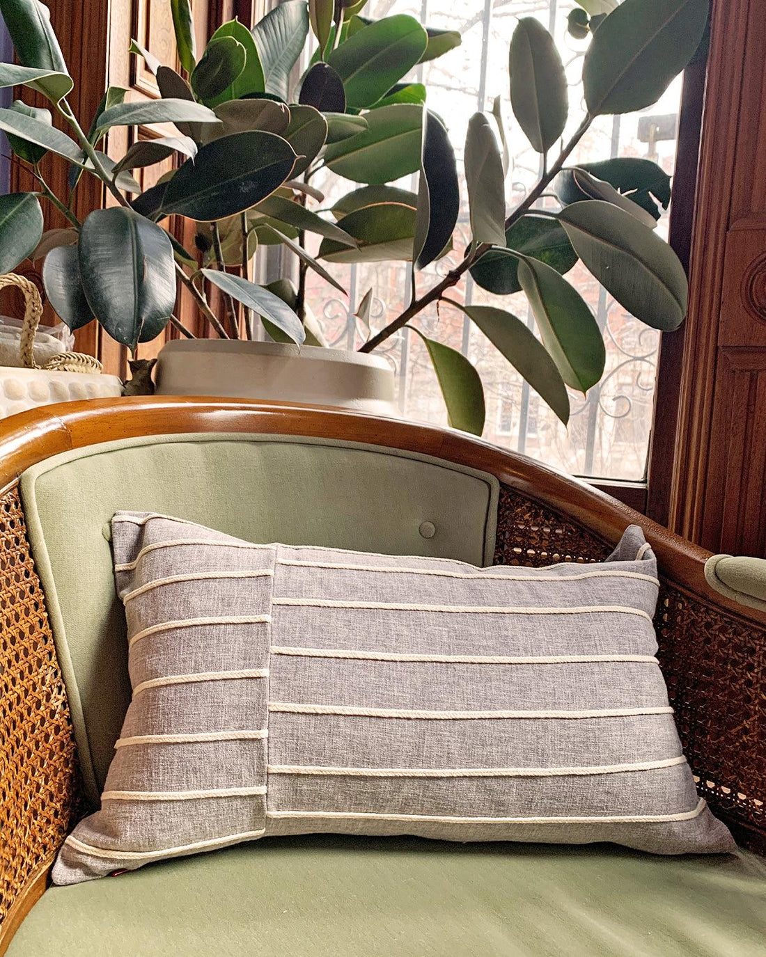 grey lumbar pillow with cream stripes on top of green wicker chair with plants in background
